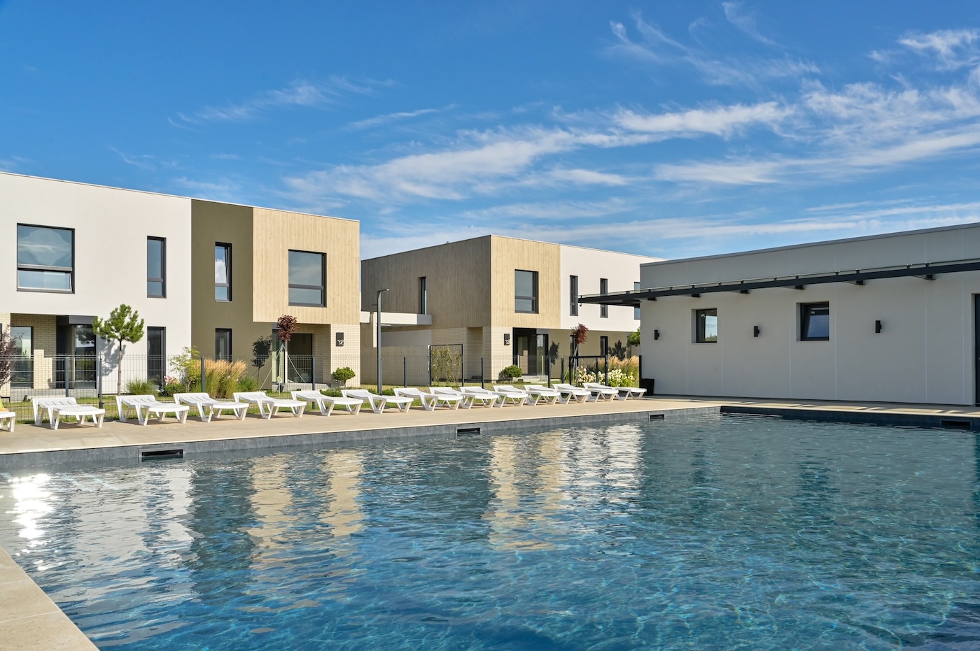 Modern apartment complex with a large outdoor pool and row of white lounge chairs under a blue sky.